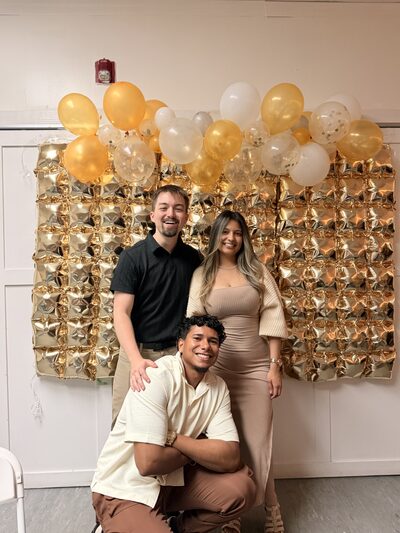 Three team members in front of a gold balloon wall at a company celebration